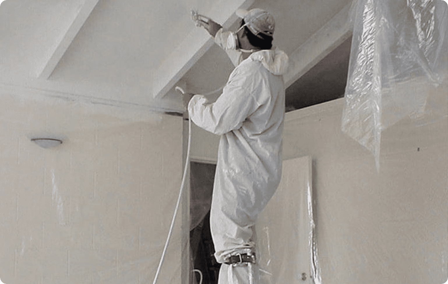 A man wearing protective wear is painting a ceiling white.